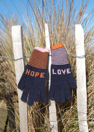 Pair of knitted gloves with 'HOPE' and 'LOVE' text hanging on a fence with grass in the background.