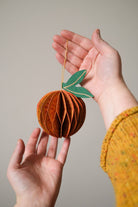 Hand holding a decorative paper fruit ornament against a neutral background