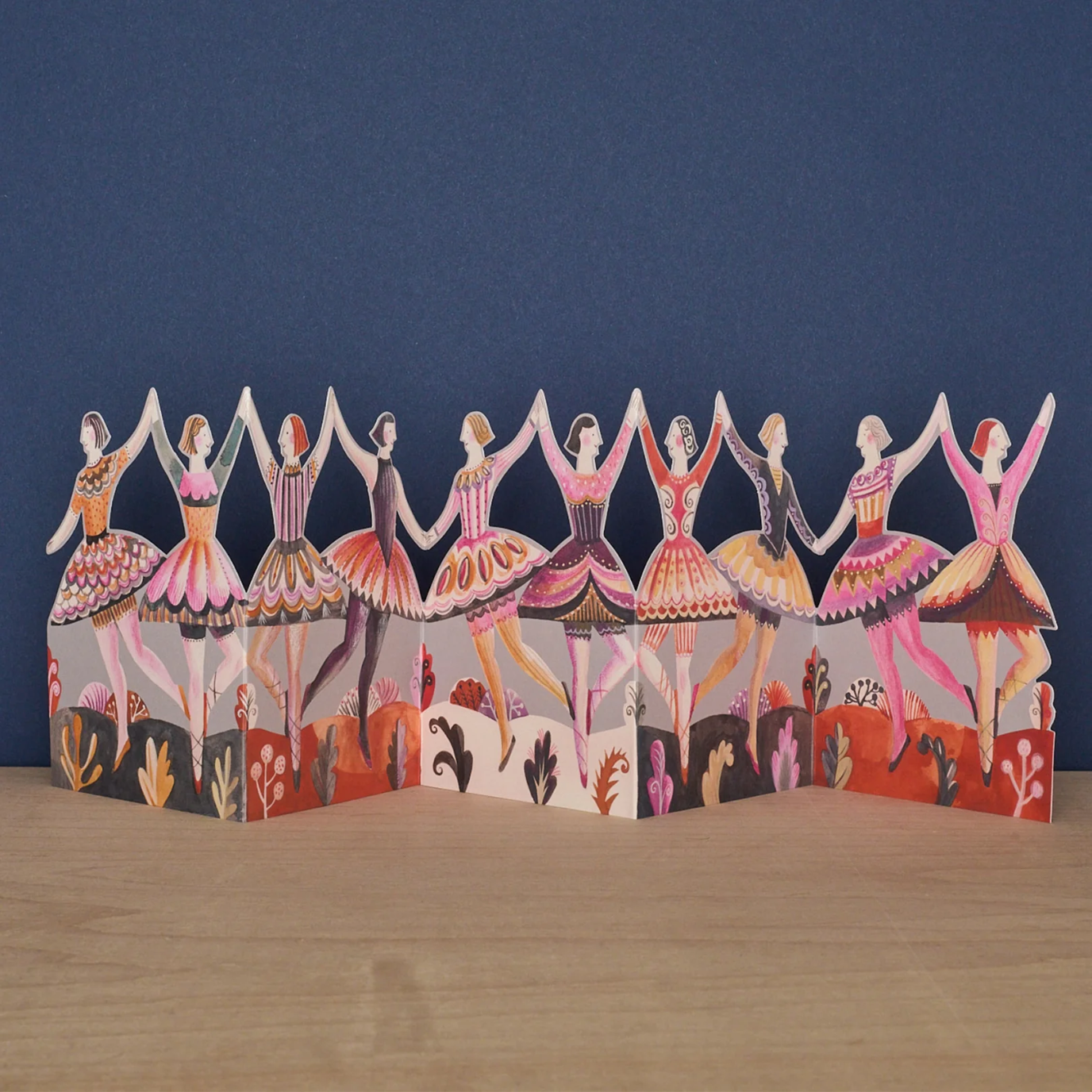 Decorative paper cutout card of ballerinas in a row on a wooden surface with a blue background