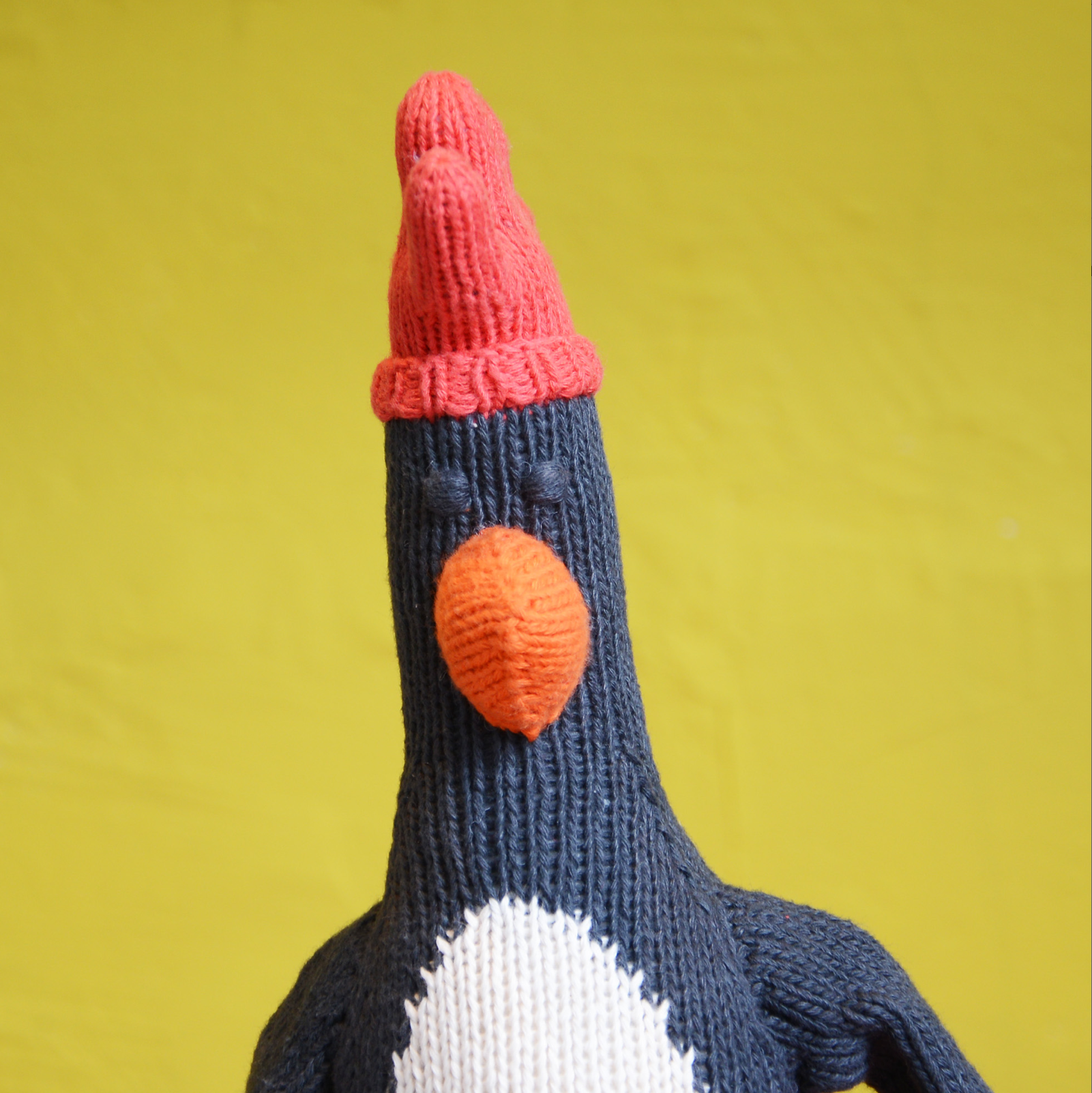 A knitted penguin toy featuring a black body, a white belly, and a bright orange beak with a red hat. The background of the photo is a solid yellow backdrop.