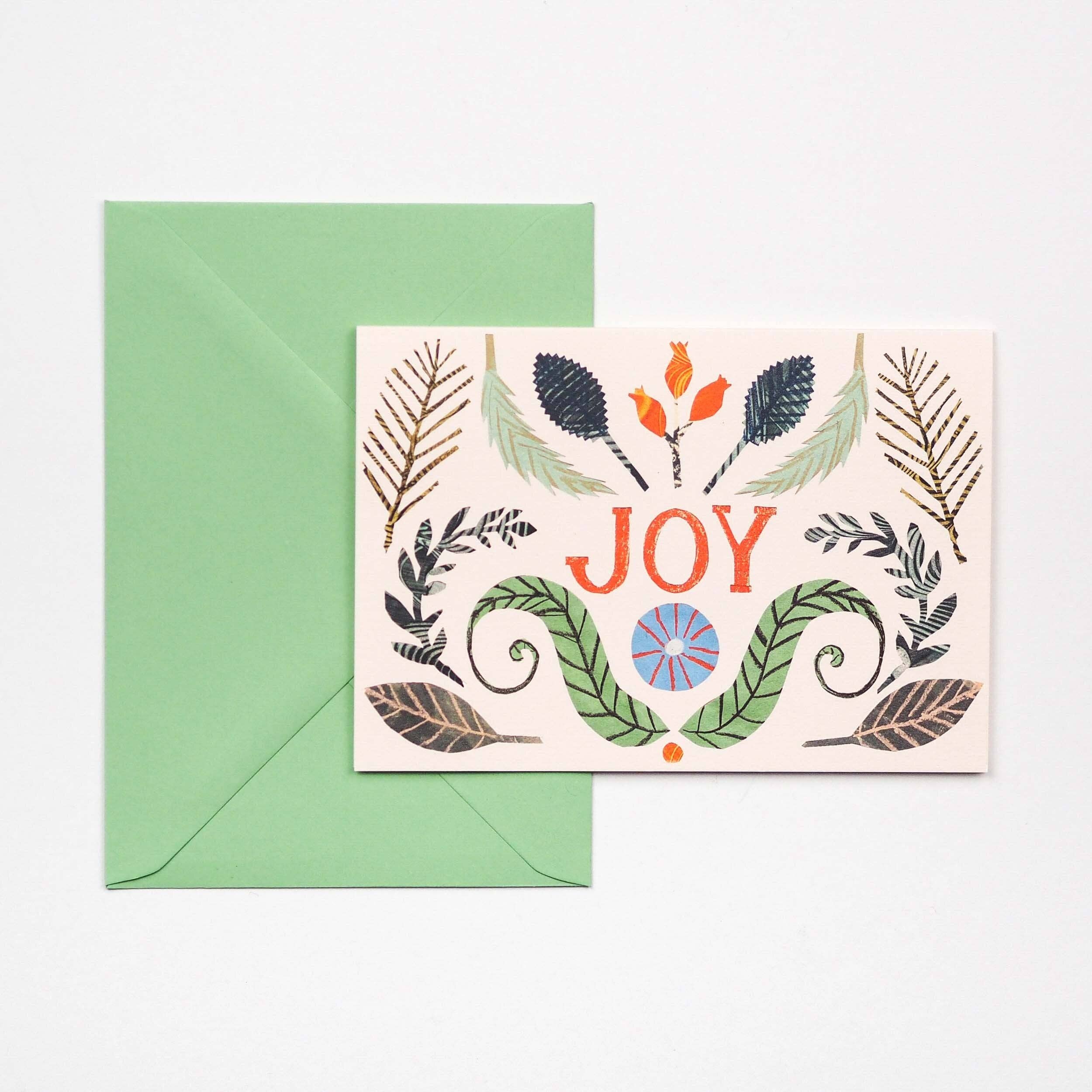 A greeting card with a botanical illustrations, placed in front of a green envelope on a light background. The card features the word “JOY” in orange-red lettering with decorative leaves and small floral motifs around it.