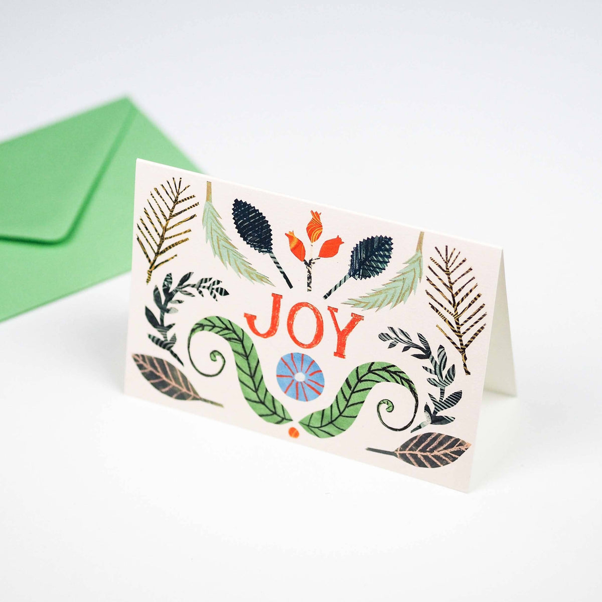 A folded greeting card standing upright on a white surface, decorated with illustrated leaves and the word “JOY” in orange-red. A green envelope sits behind the card.