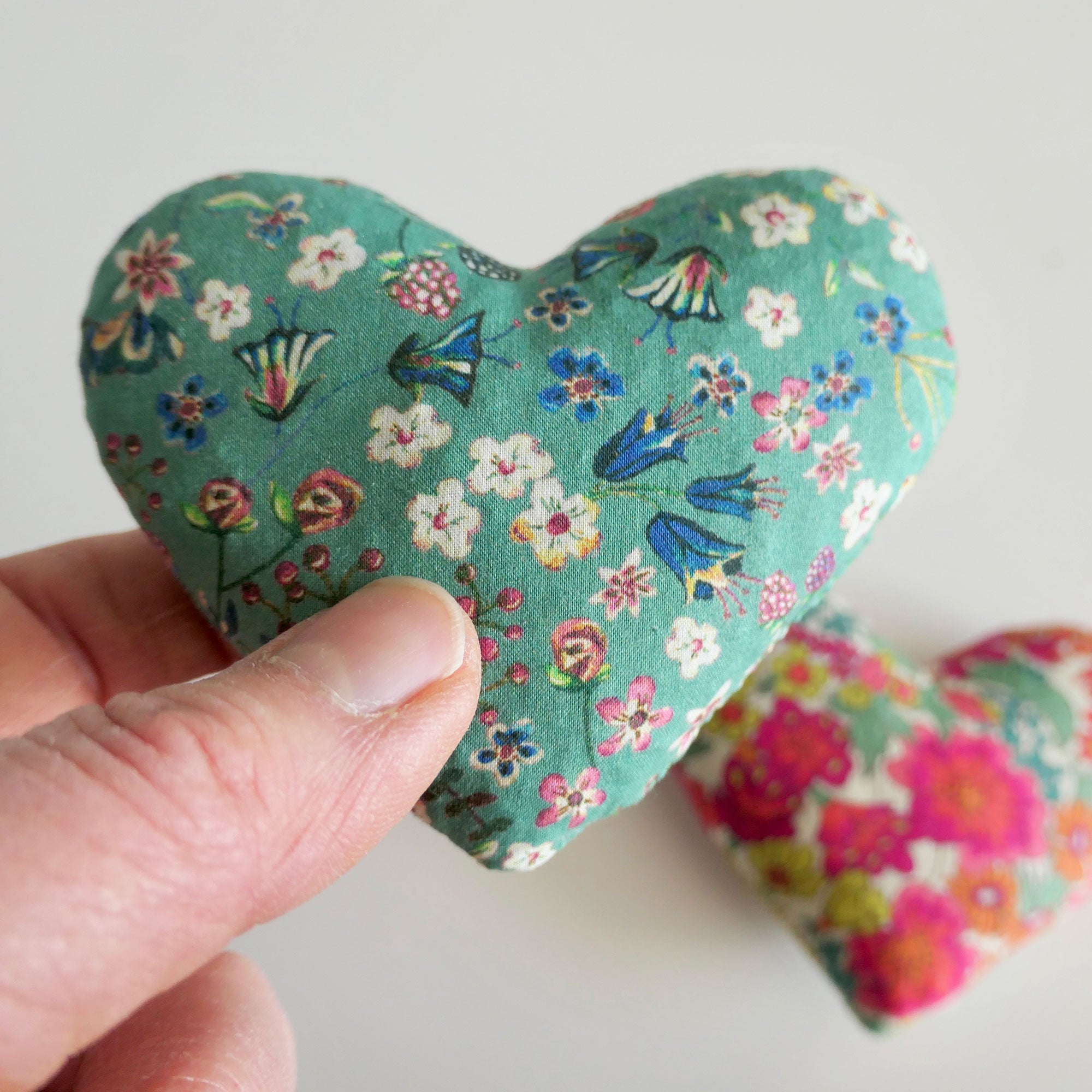 Floral-patterned heart-shaped fabric item held by a hand on a light background