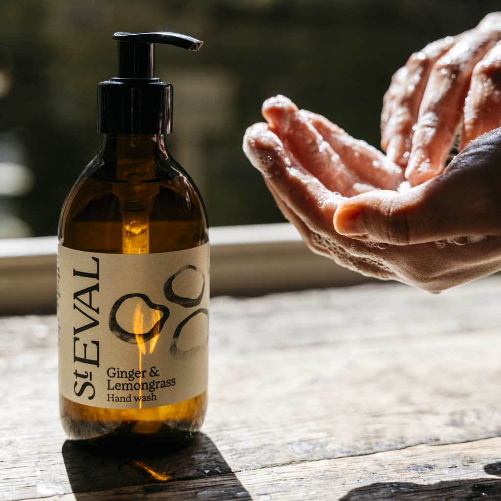A bottle of St Eval Ginger & Lemongrass hand soap sits on a wooden windowsill. A pair of hands covered in soapy suds can be seen in shot.