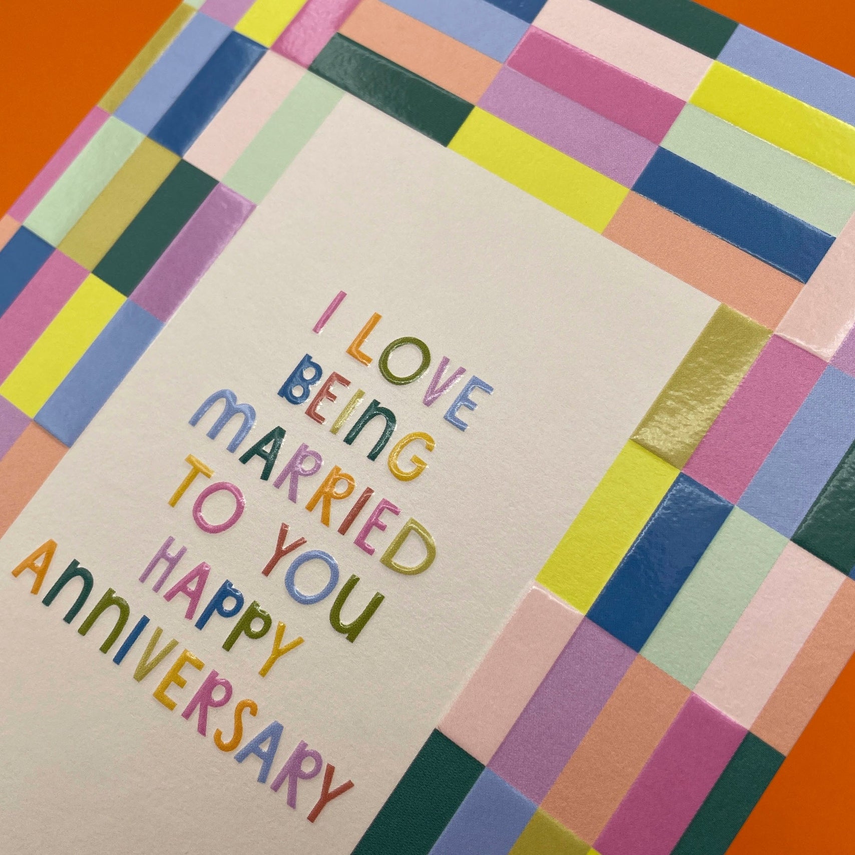 Bold and colourful anniversary card. Reads “I LOVE BEING MARRIED TO YOU HAPPY ANNIVERSARY”. With a colourful block pattern border. Detail close up.