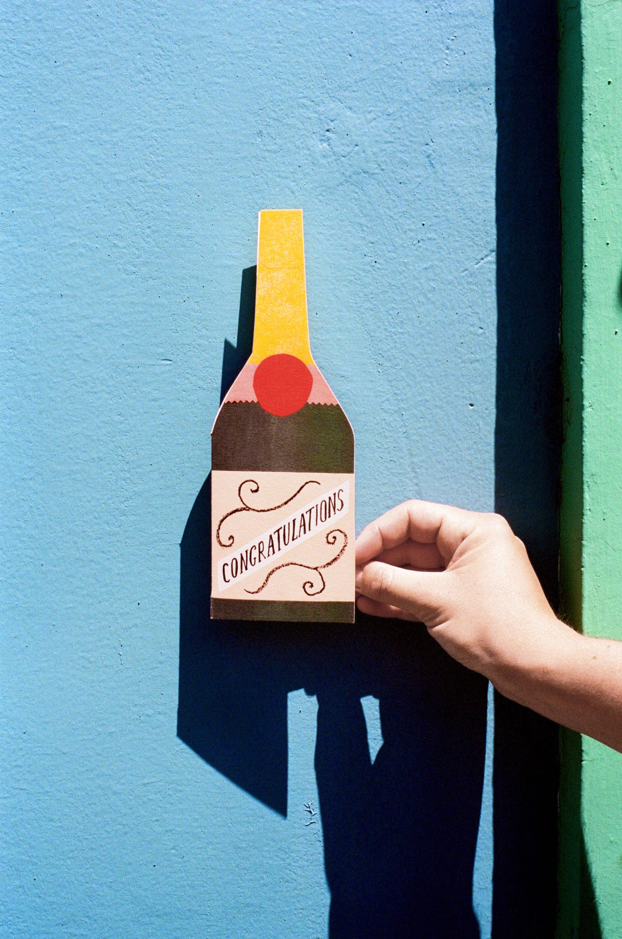Hand holding a bottle-shaped 'Congratulations' card against a blue wall.
