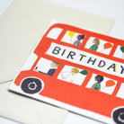 Birthday card with a red bus design featuring illustrations of people and balloons on a beige background.