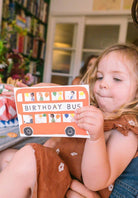 Child holding a card with 'Birthday Bus' text in a cozy indoor setting.