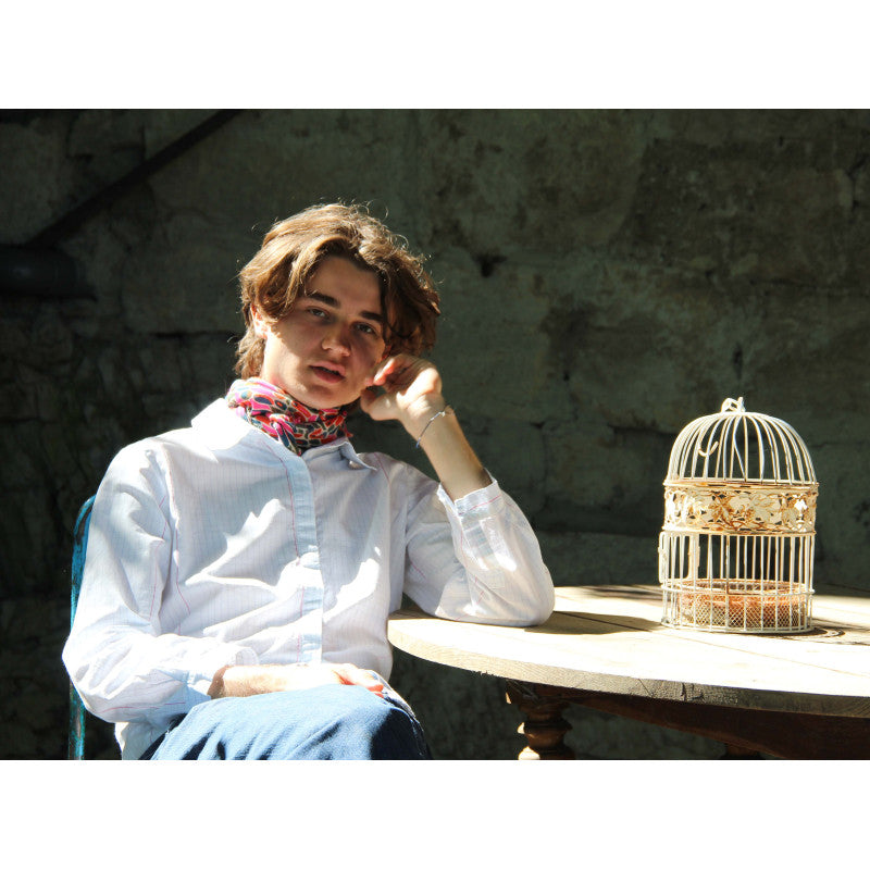A young man with light brown hair sits at a wooden table wearing a white button-down shirt and a colorful patterned neck scarf. A small white birdcage sits on the table next to him against a rustic stone wall background.