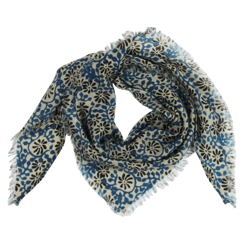 A square scarf with fringed edges, featuring a blue and black floral block-print pattern on a cream background, arranged in a loose knot on a light gray studio background.