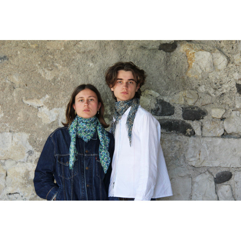 Two models standing in front of a rustic stone wall, both wearing matching blue and green patterned neck scarves.