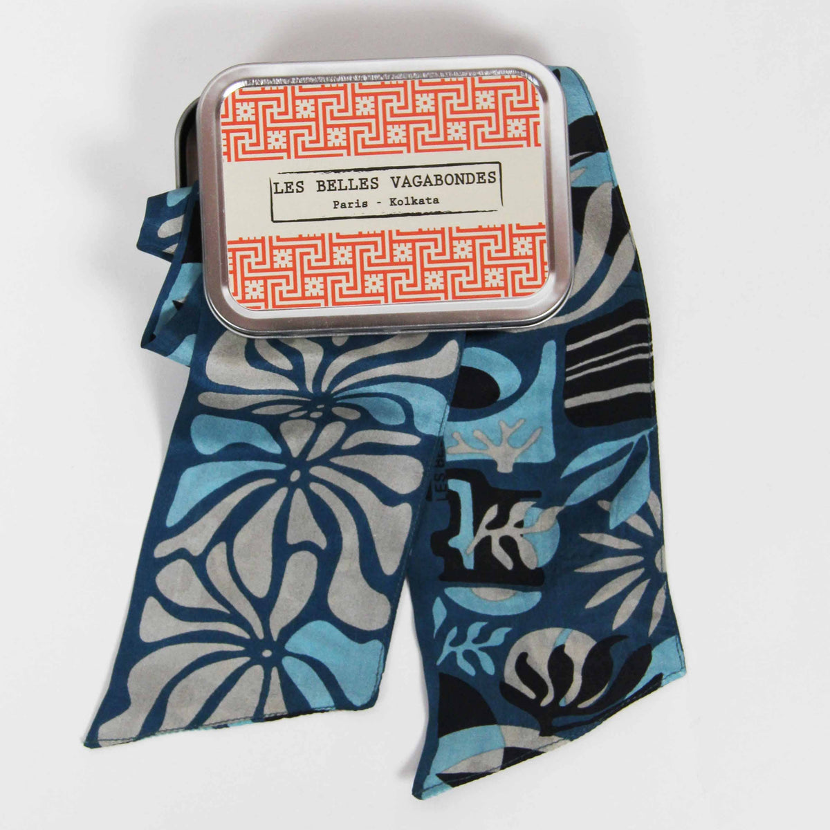 A patterned silk scarf in shades of blue, gray, and black, draped next to its silver metal tin packaging with a red and white label r