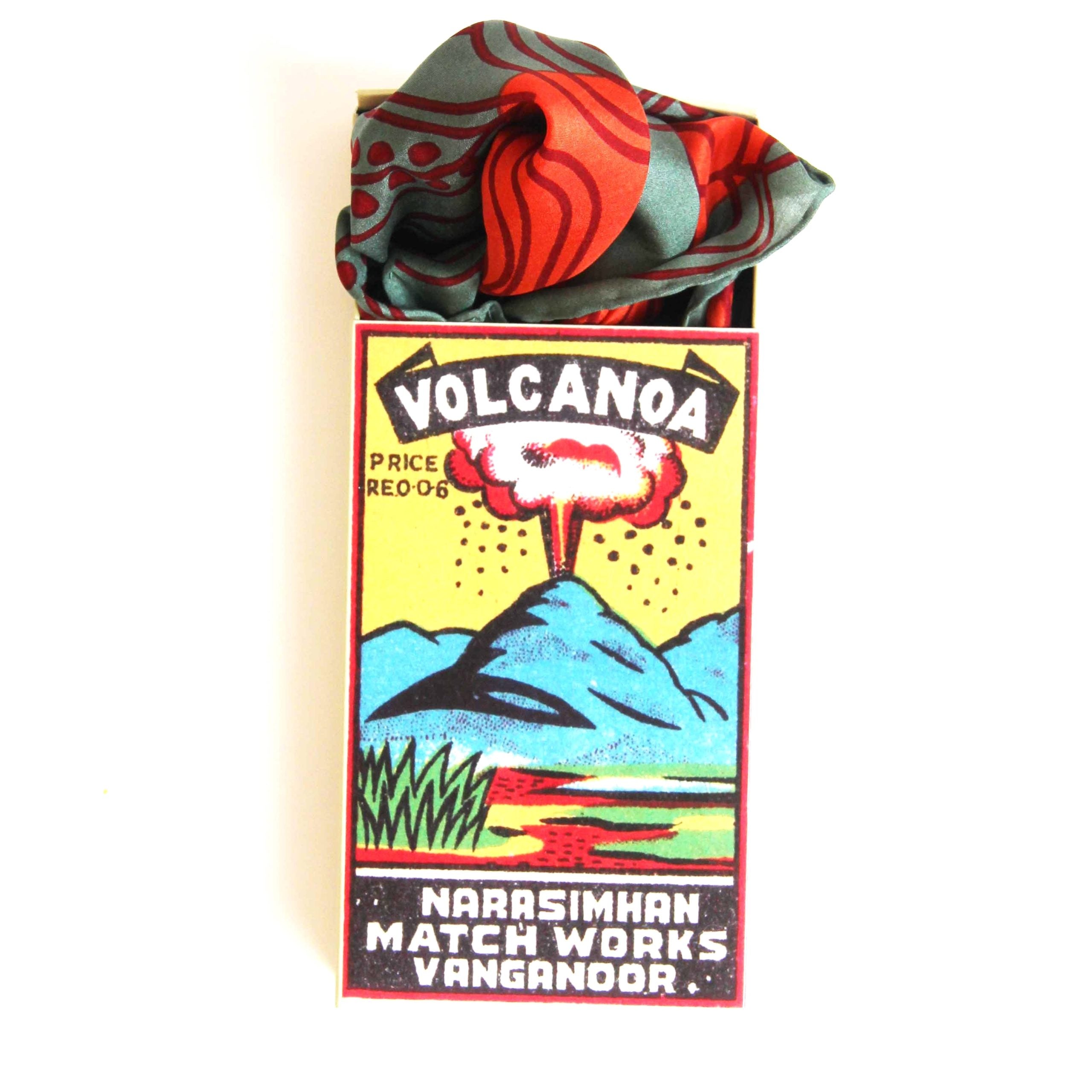 Matchbox with a colorful design featuring a volcano and text, containing a red and green patterned fabric.