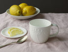 White mug with embossed design, plate of lemons, and spoon on a pink cloth.