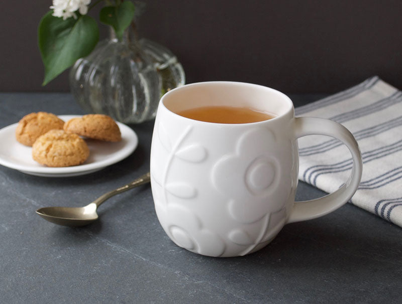 White textured mug with tea on a dark surface with cookies and a vase in the background