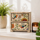 Decorative wooden box with floral and bird patterns on a white surface with a candle and plant in the background.