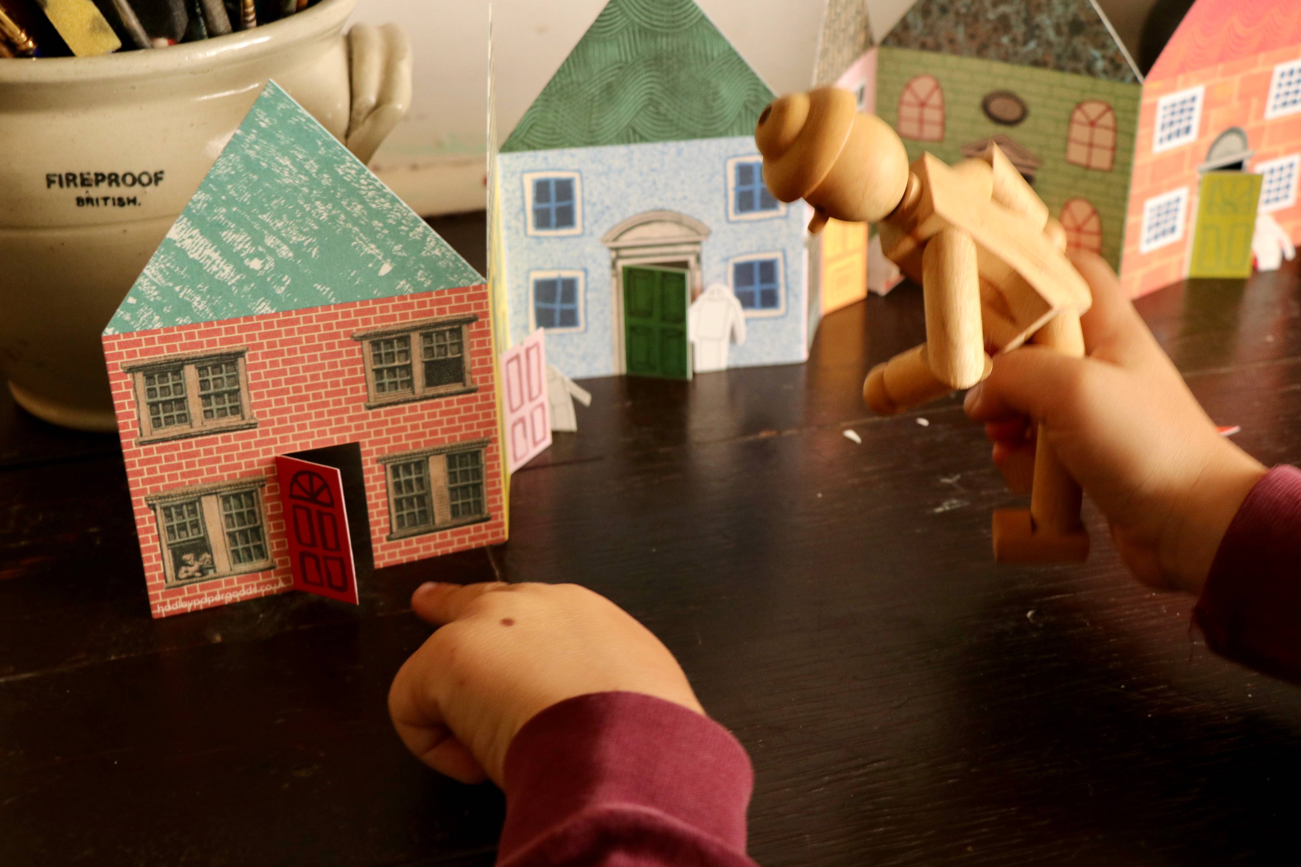 Children playing with wooden toy figures and paper house models on a table.
