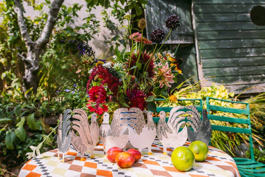 Decorative paper chickens on a table with apples and limes, set against a garden backdrop.