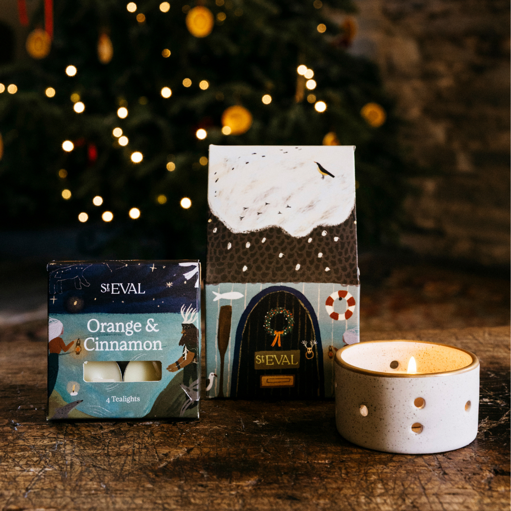 Two candle boxes and a lit candle on a wooden surface with a Christmas tree in the background.