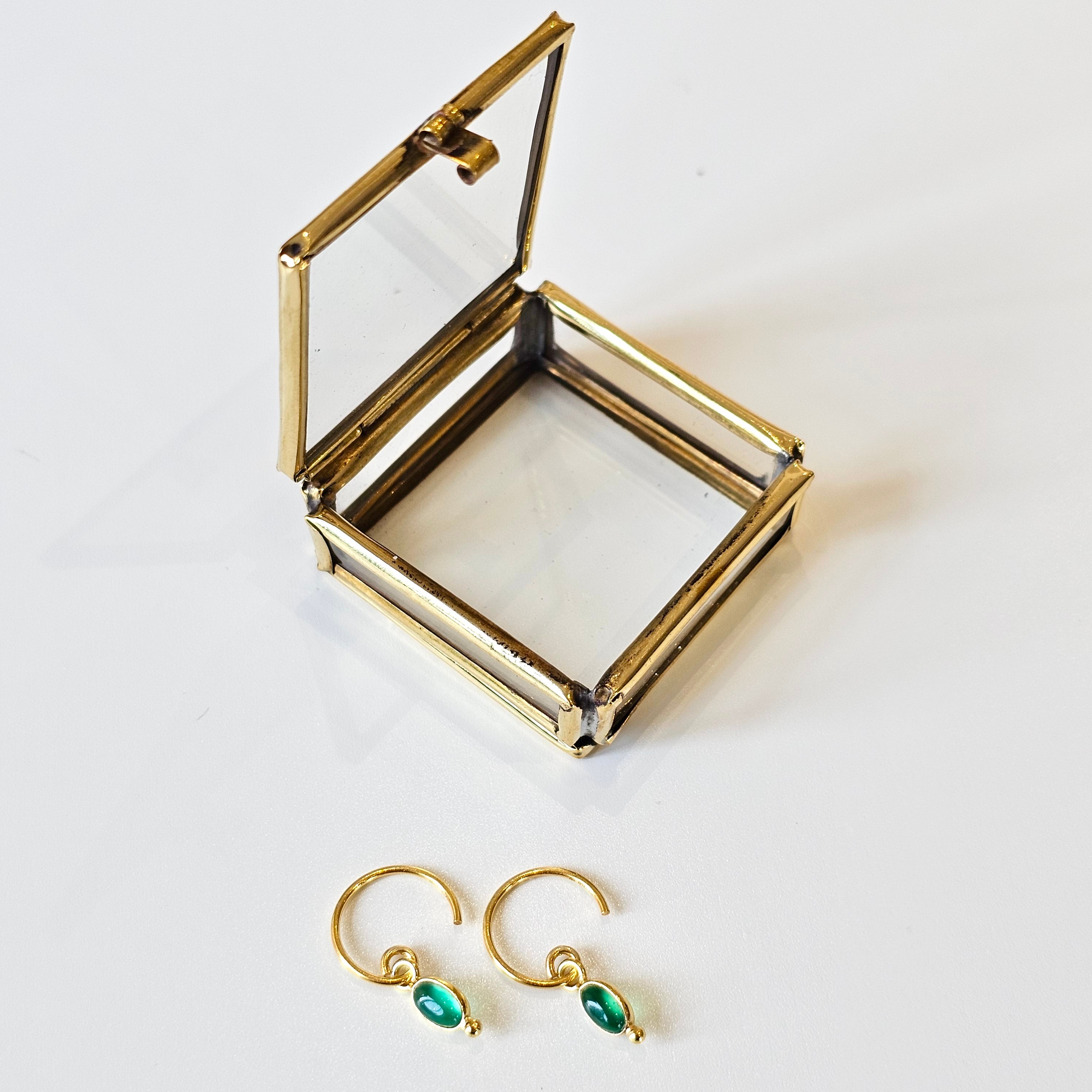 Gold earrings with green gemstones on a white surface next to an open gold jewelry box.