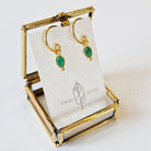 Gold hoop earrings with green stones on a branded card in a gold stand.