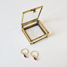 Gold earrings with amethyst stones next to an open gold jewellery box on a white background