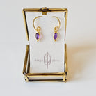 Gold earrings with amethyst stones in a gold display case on a white background