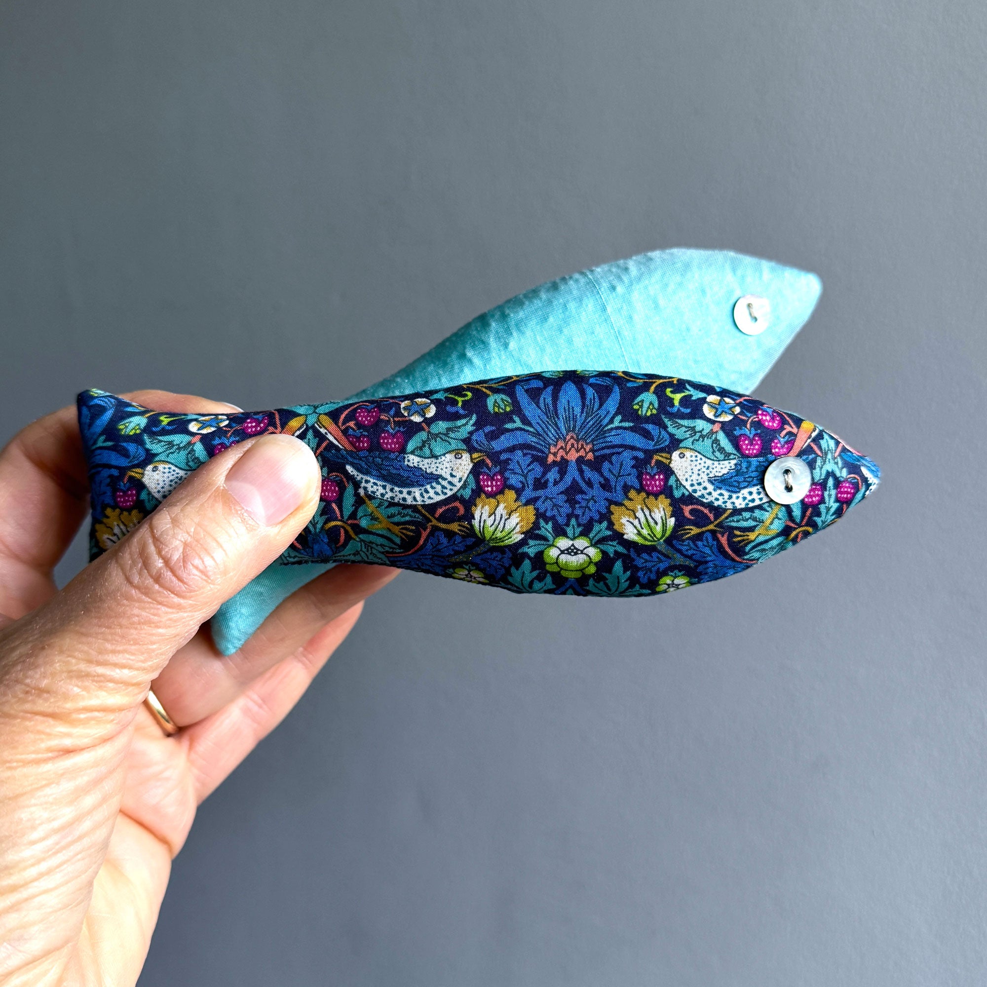 Hand holding a fish-shaped sachet with Strawberry Thief pattern against a grey background