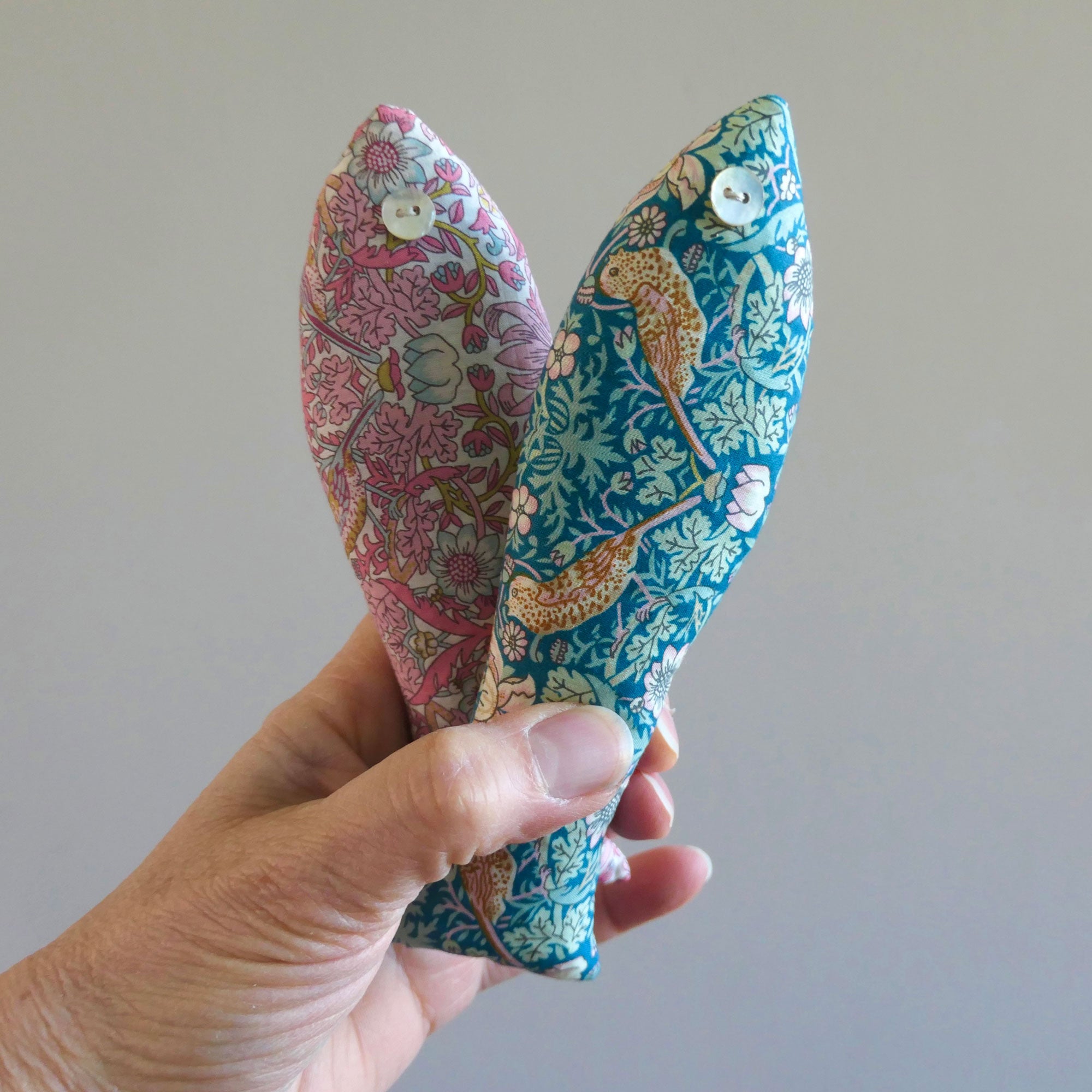 Hand holding two floral-patterned fish-shaped  sachets in Strawberry Thief Liberty Fabric against a plain background