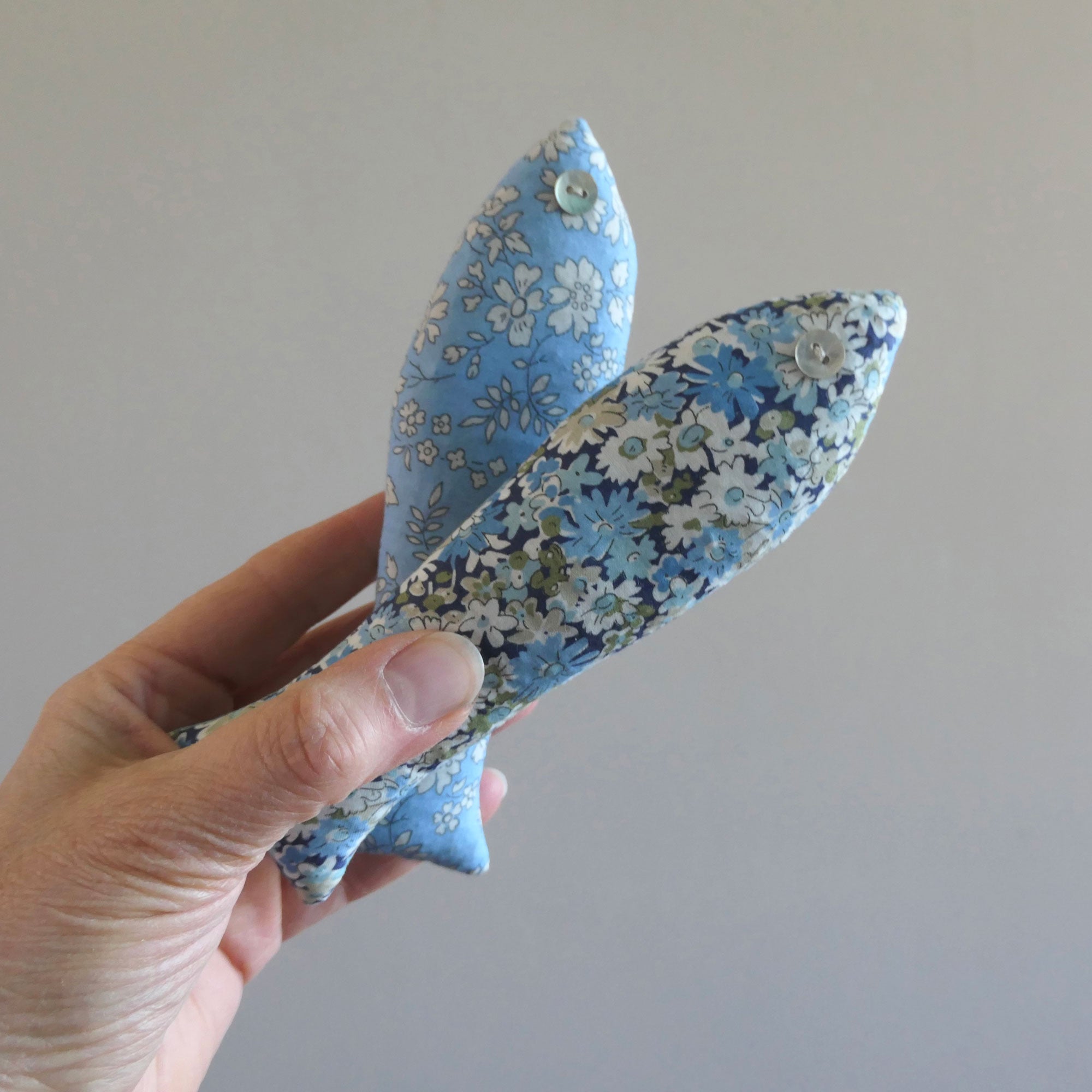 Hand holding two blue floral fabric fish-shaped sachets against a plain background