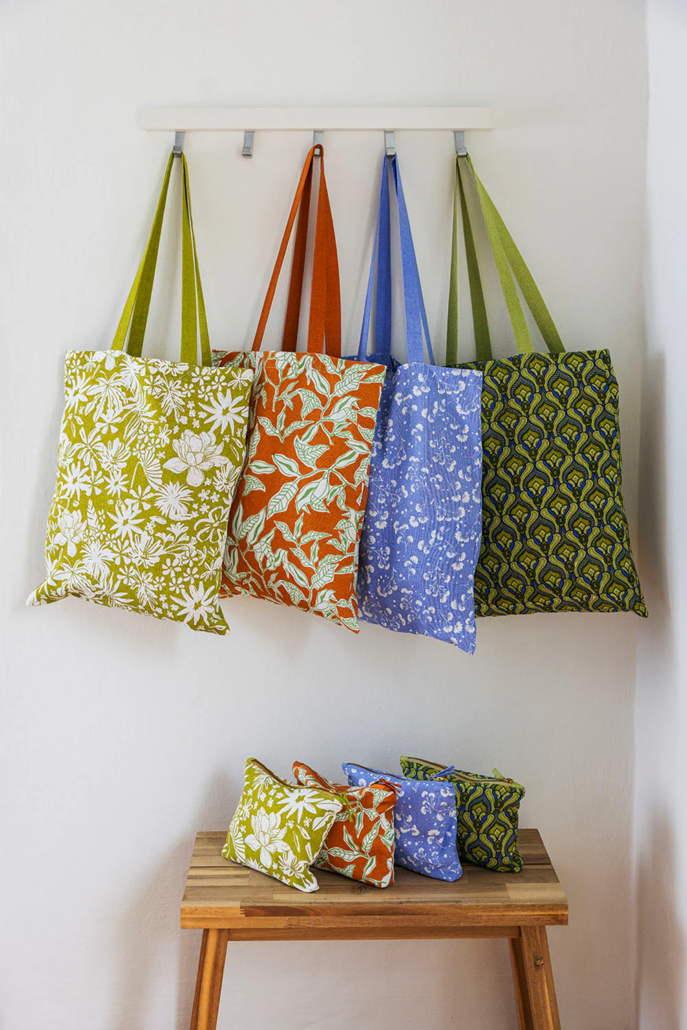 Colourful patterned bags hanging on a wall rack with additional bags on a wooden stool below.