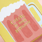 Greetings card featuring a pint mug of beer on a mustard yellow background. Reads “Cheers to your birthday” in gold embossing. Detail close up.