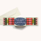 A Christmas greeting card set with a central card reading 'MERRY CHRISTMAS!' in white text on a blue oval, flanked with pink, red, and green geometric designs, shown with an off-white envelope on a light beige background.