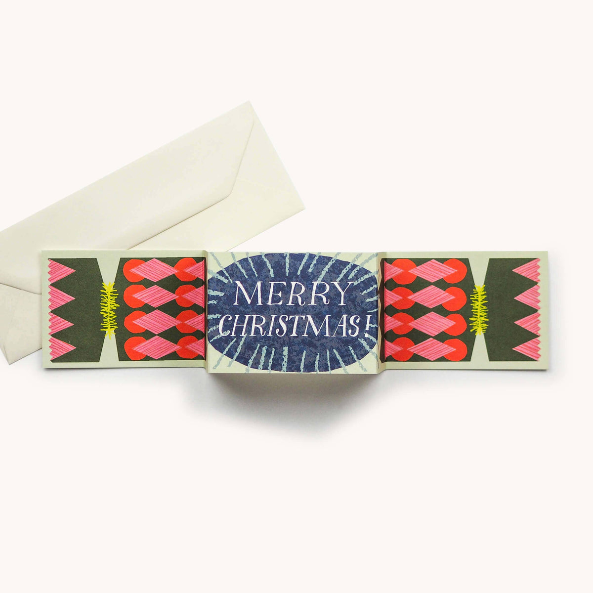 A Christmas greeting card set with a central card reading 'MERRY CHRISTMAS!' in white text on a blue oval, flanked with pink, red, and green geometric designs, shown with an off-white envelope on a light beige background.