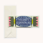 A Christmas card designed like a cracker, shown folded flat with a cream envelope behind it on a light background. The center of the card is blue with white radial lines and the words “MERRY CHRISTMAS!” in white.