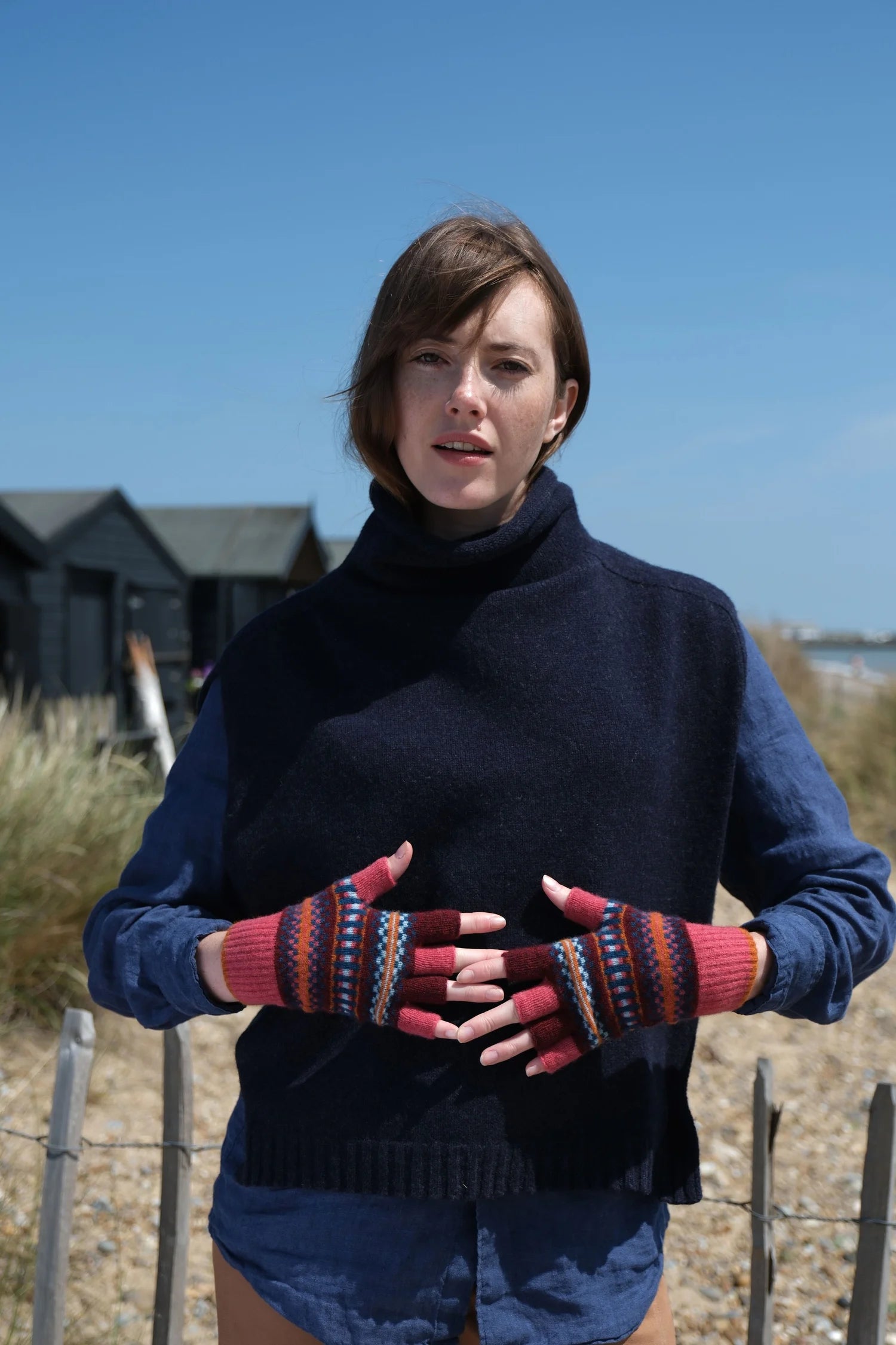 Woman wearing a blue sweater and colorful fingerless gloves on a beach with huts in the background.