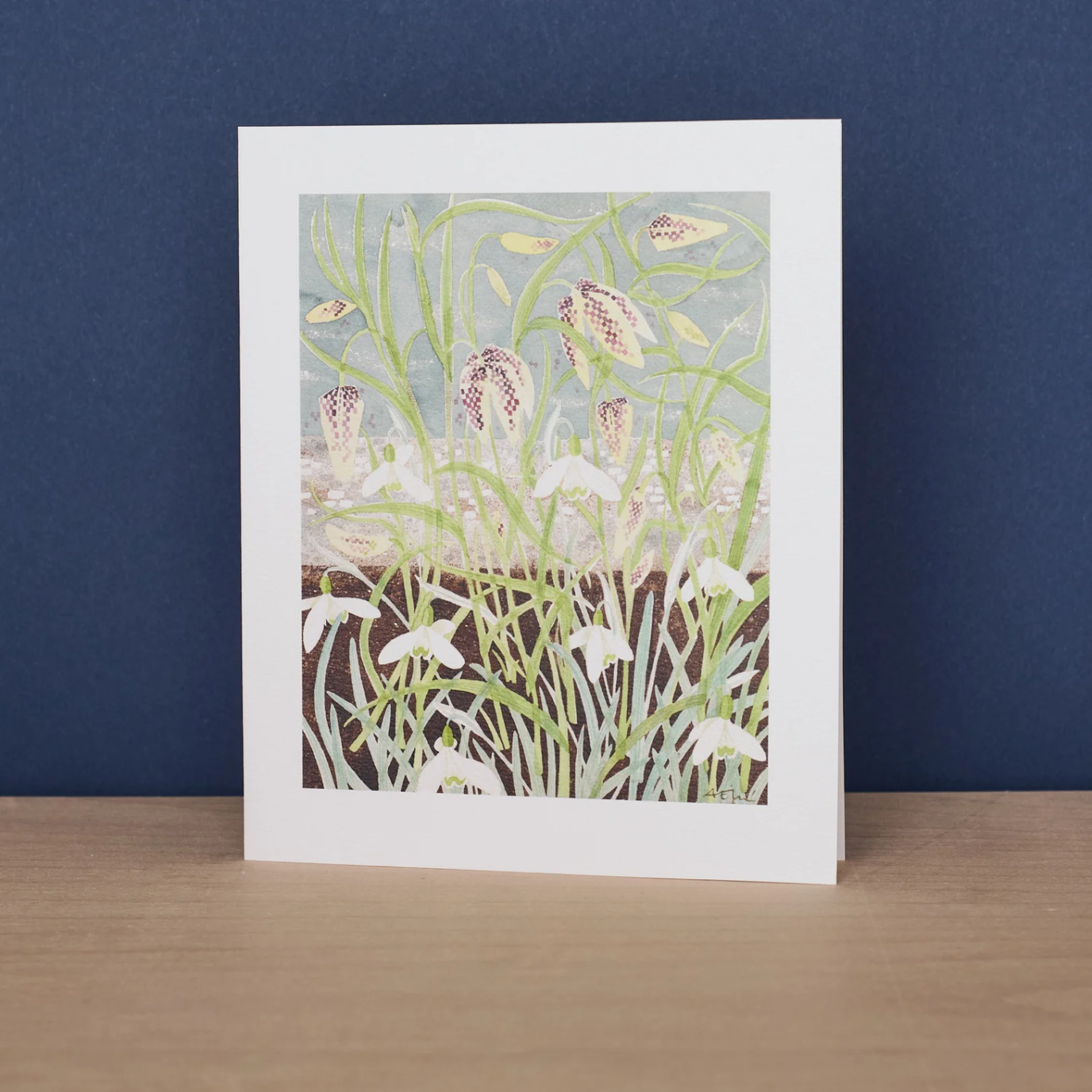 Greetings card featuring artwork of fritillaries and snowdrops. Card sits against a blue background on a wooden surface.