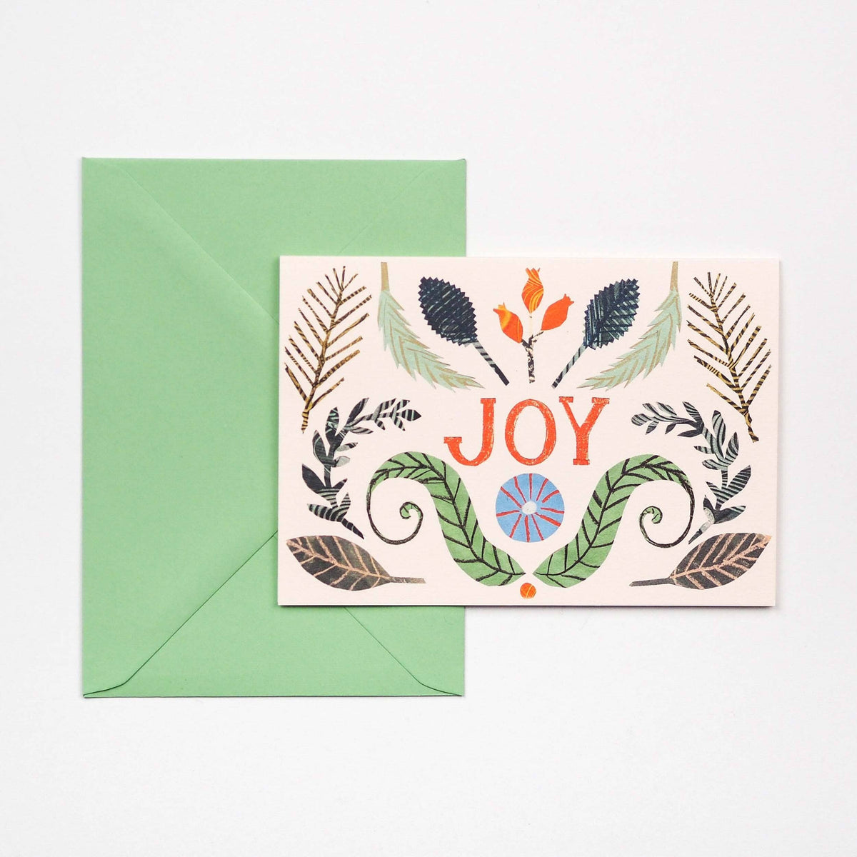 A greeting card with a  botanical illustrations, placed in front of a green envelope on a light background. The card features the word “JOY” in orange-red lettering with decorative leaves and small floral motifs around it.