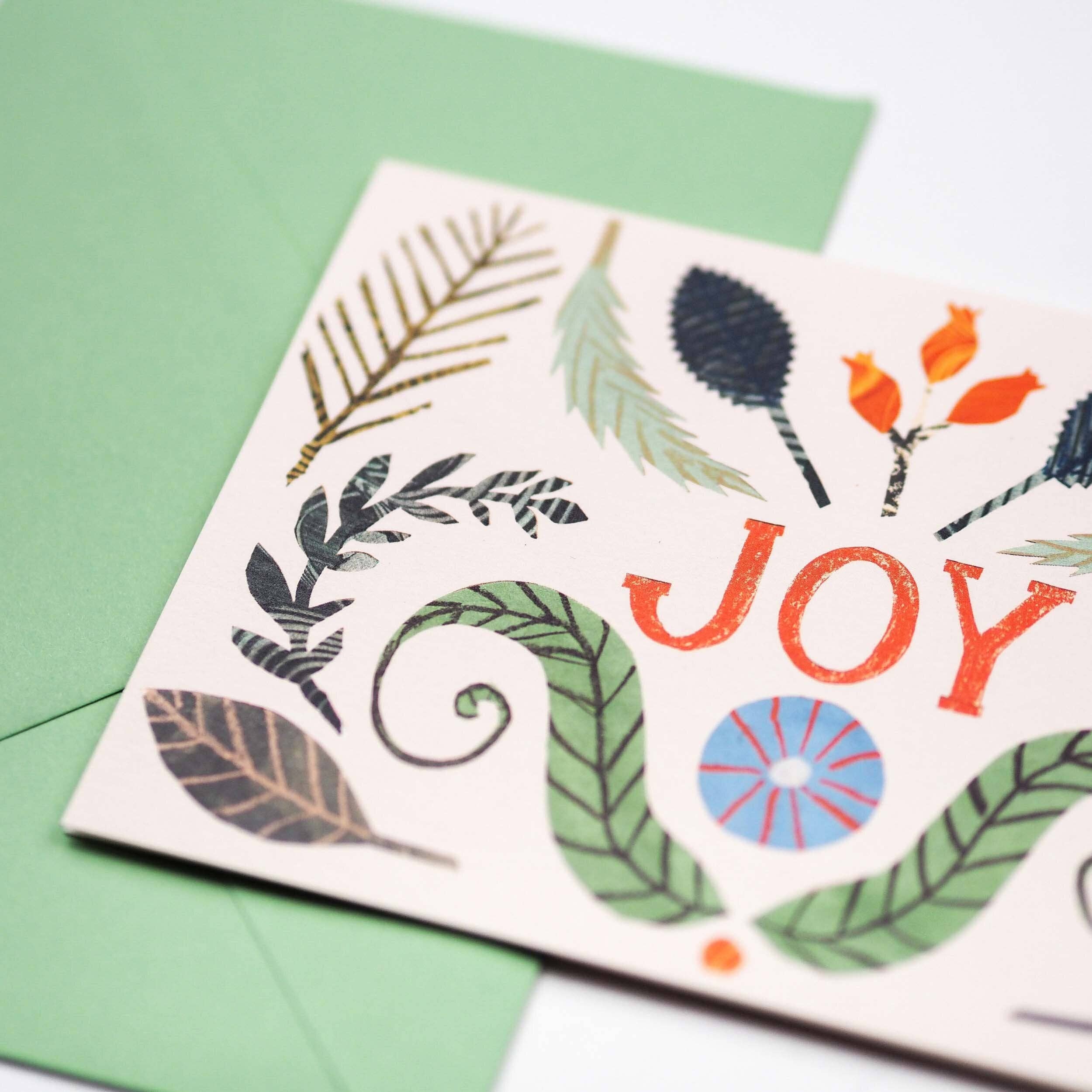 A close-up of a printed greeting card with the word “JOY” in orange-red, decorated with illustrated leaves and floral motifs in green, blue, orange, and black, layered over a light green envelope on a white background.
