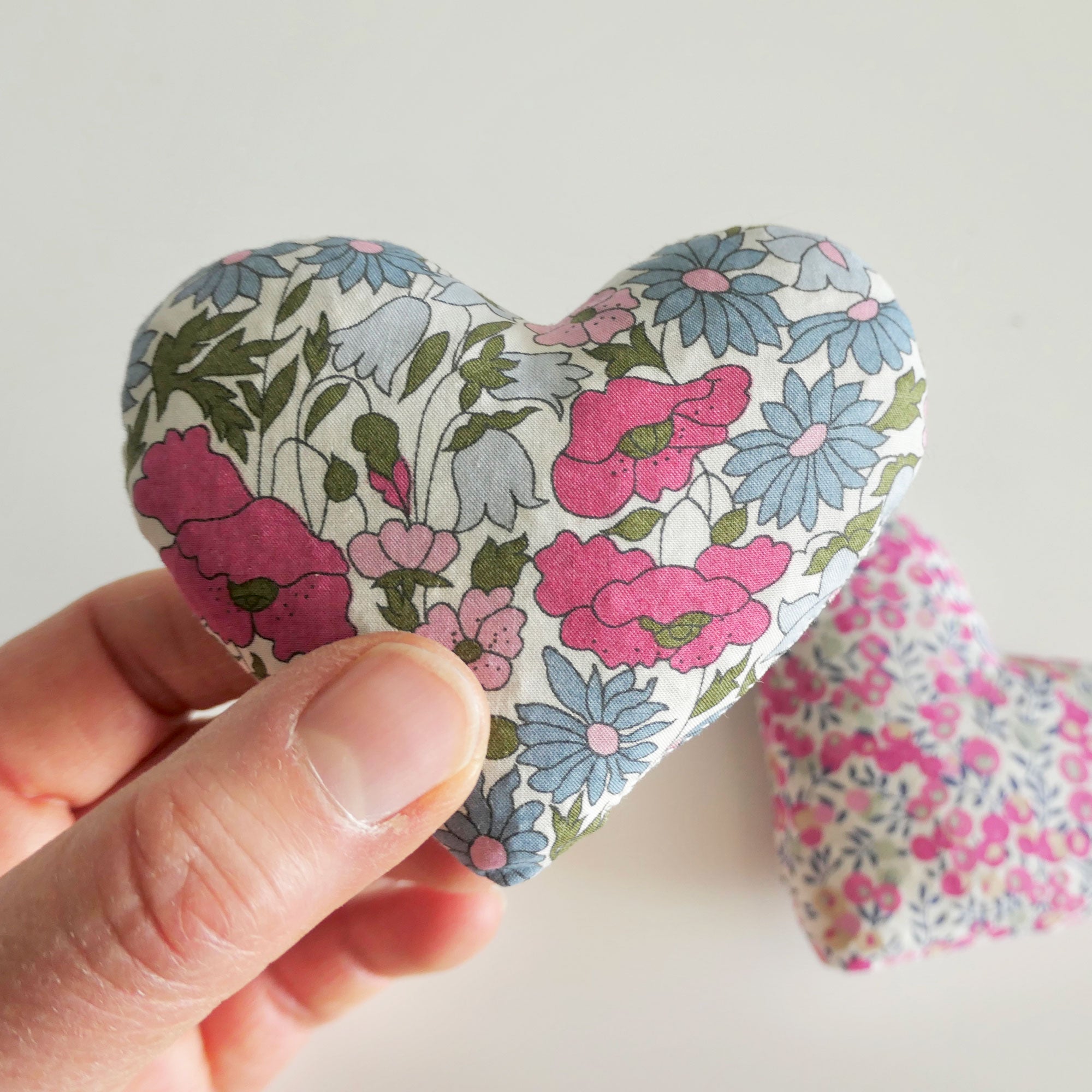 Floral-patterned heart-shaped fabric item held by a hand on a light background