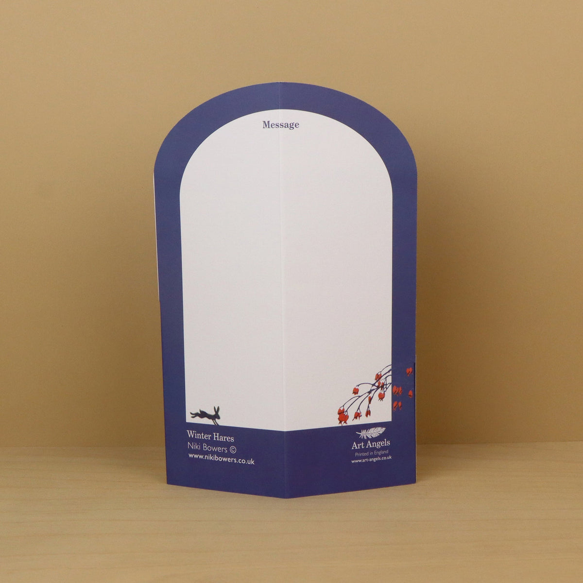 A freestanding arched paper card  with a white writing area and a dark blue border, placed on a light wood surface against a tan background. The bottom section includes printed branding and contact details.