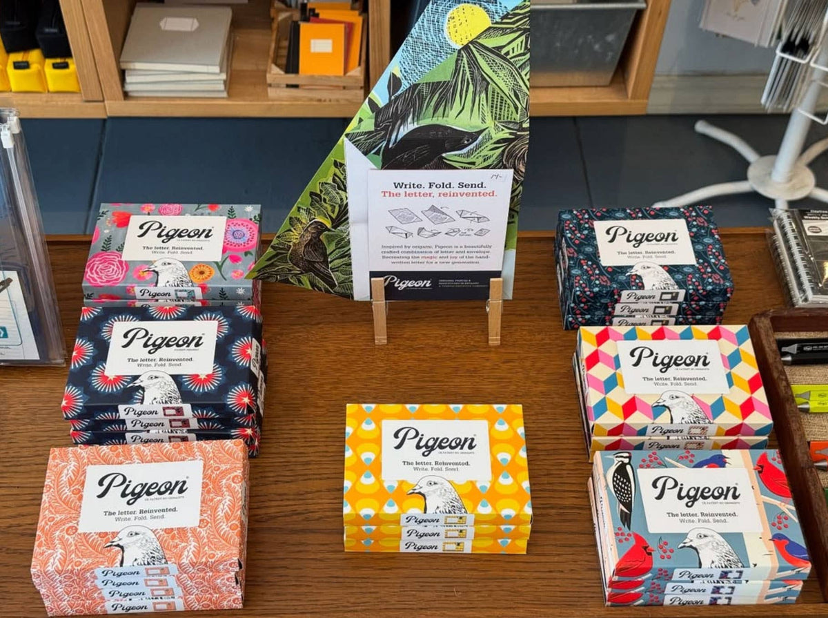 Collection of Pigeon product boxes on a wooden surface with a colourful poster in the background.