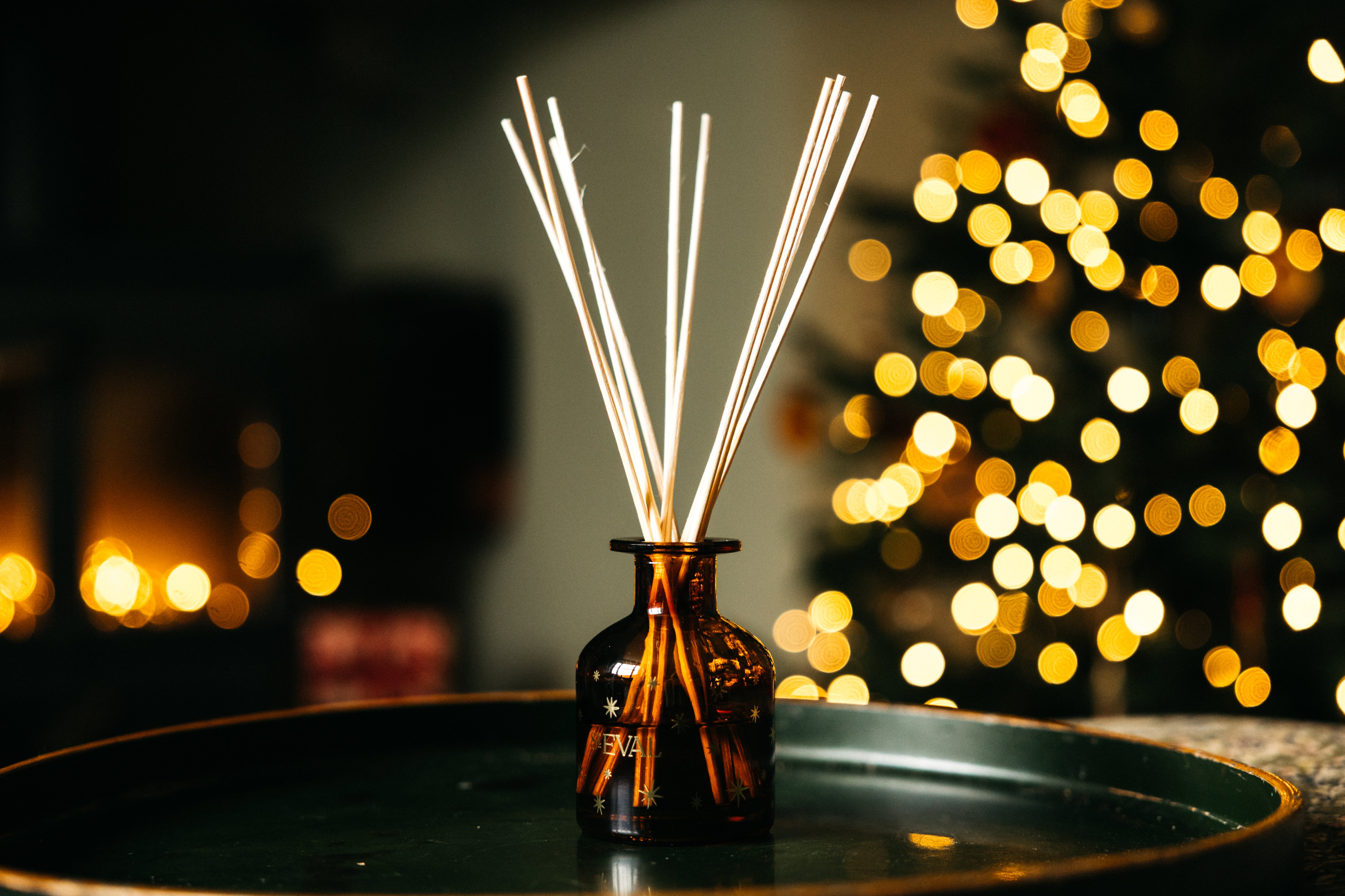 A reed diffuser with a small amber glass bottle and several natural reed sticks, set on a dark tray with a warm, blurred holiday tree of lights in the background.