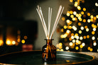 A reed diffuser with a small amber glass bottle and several natural reed sticks, set on a dark tray with a warm, blurred holiday tree of lights in the background.