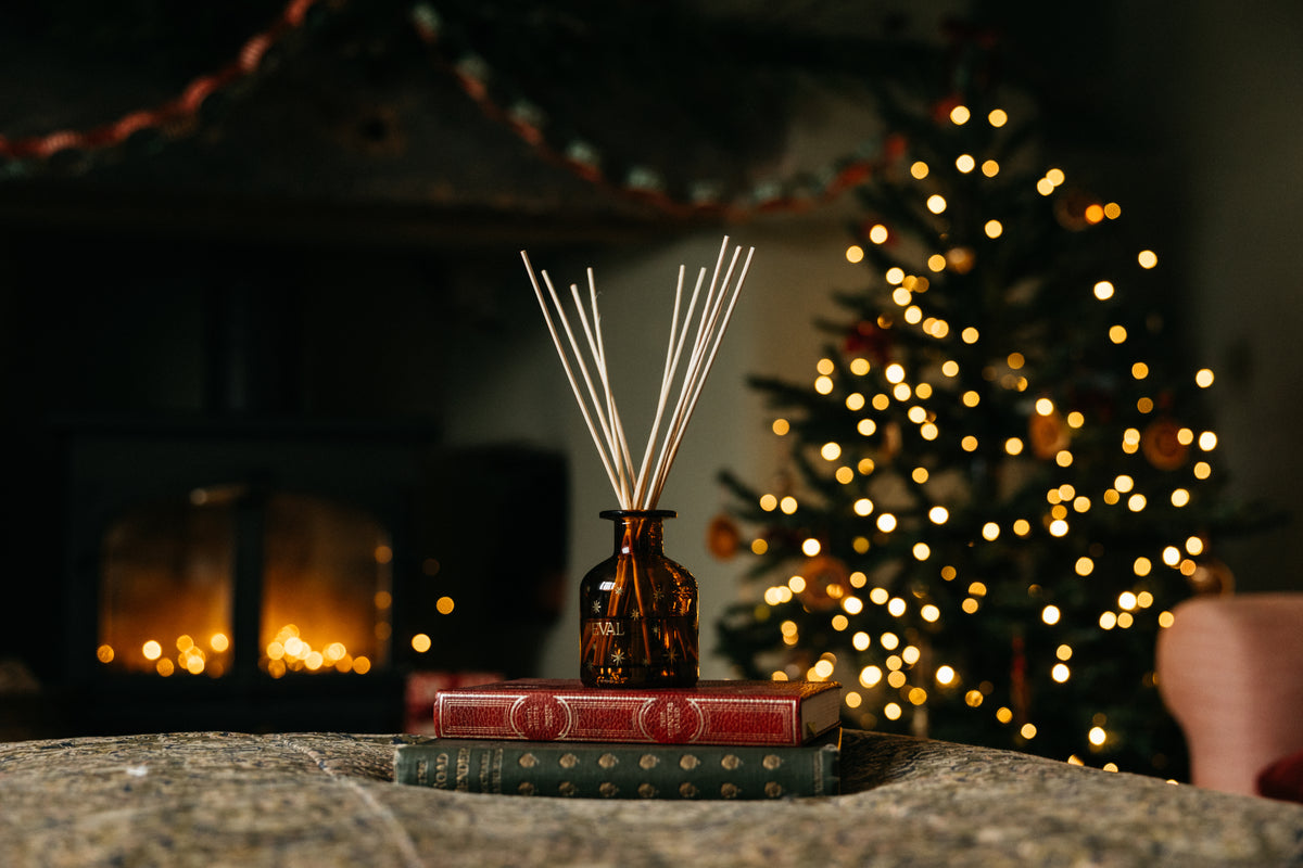 A reed diffuser in an amber glass bottle sits on two stacked vintage-style books on a stone surface, with a blurred fireplace and a glowing Christmas tree in the background.