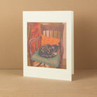 A white matted art print card featuring a painting of a black cat curled up on a wooden chair with an orange pillow, set against a warm beige background.