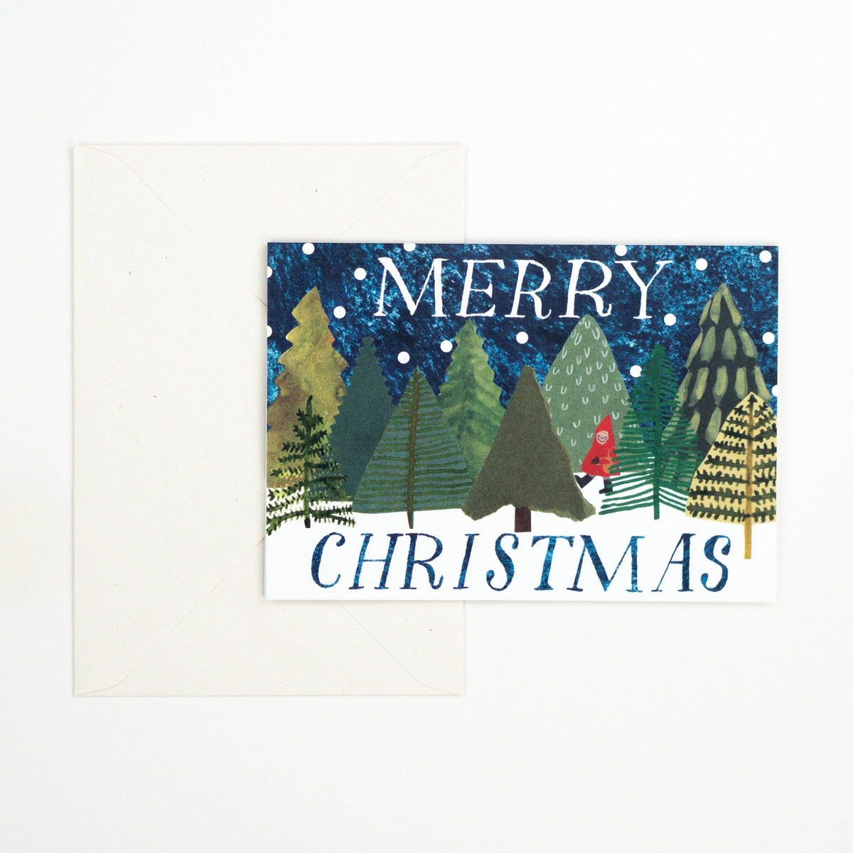 A Christmas greeting card with a white envelope behind it. The card features an illustrated winter forest scene with pine trees, snow dots, and the words “MERRY CHRISTMAS” in white and blue lettering.