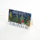 A folded Christmas greeting card standing upright on a white surface. The card features a winter forest scene with pine trees, snow, and the words “MERRY CHRISTMAS” in blue lettering.