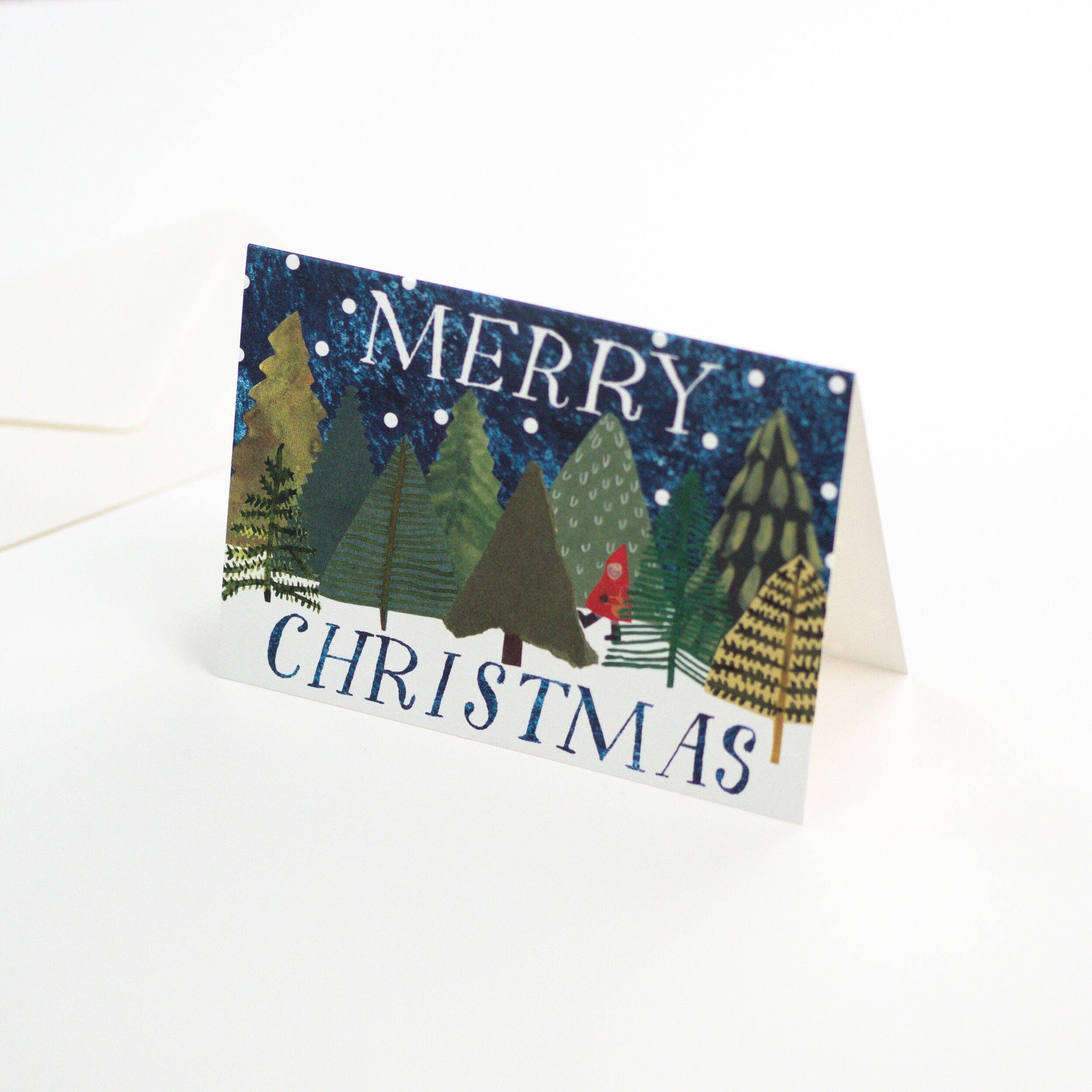 A folded Christmas greeting card standing upright on a white surface. The card features a winter forest scene with pine trees, snow, and the words “MERRY CHRISTMAS” in blue lettering.