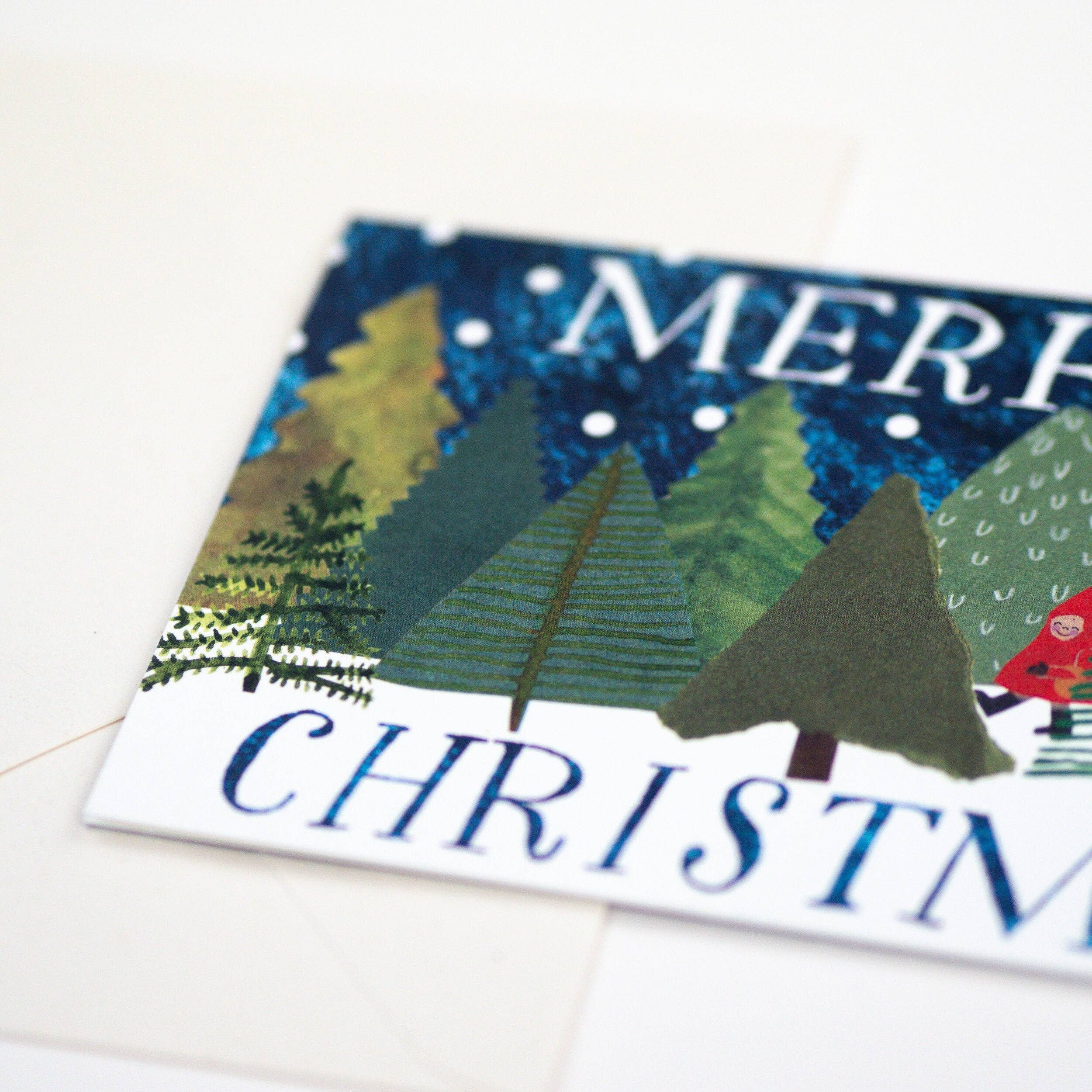 A close-up of a Christmas greeting card featuring a textured, collage-style winter forest with green and yellow trees under a dark blue sky, with the words 'MERRY CHRISTMAS' partially visible.